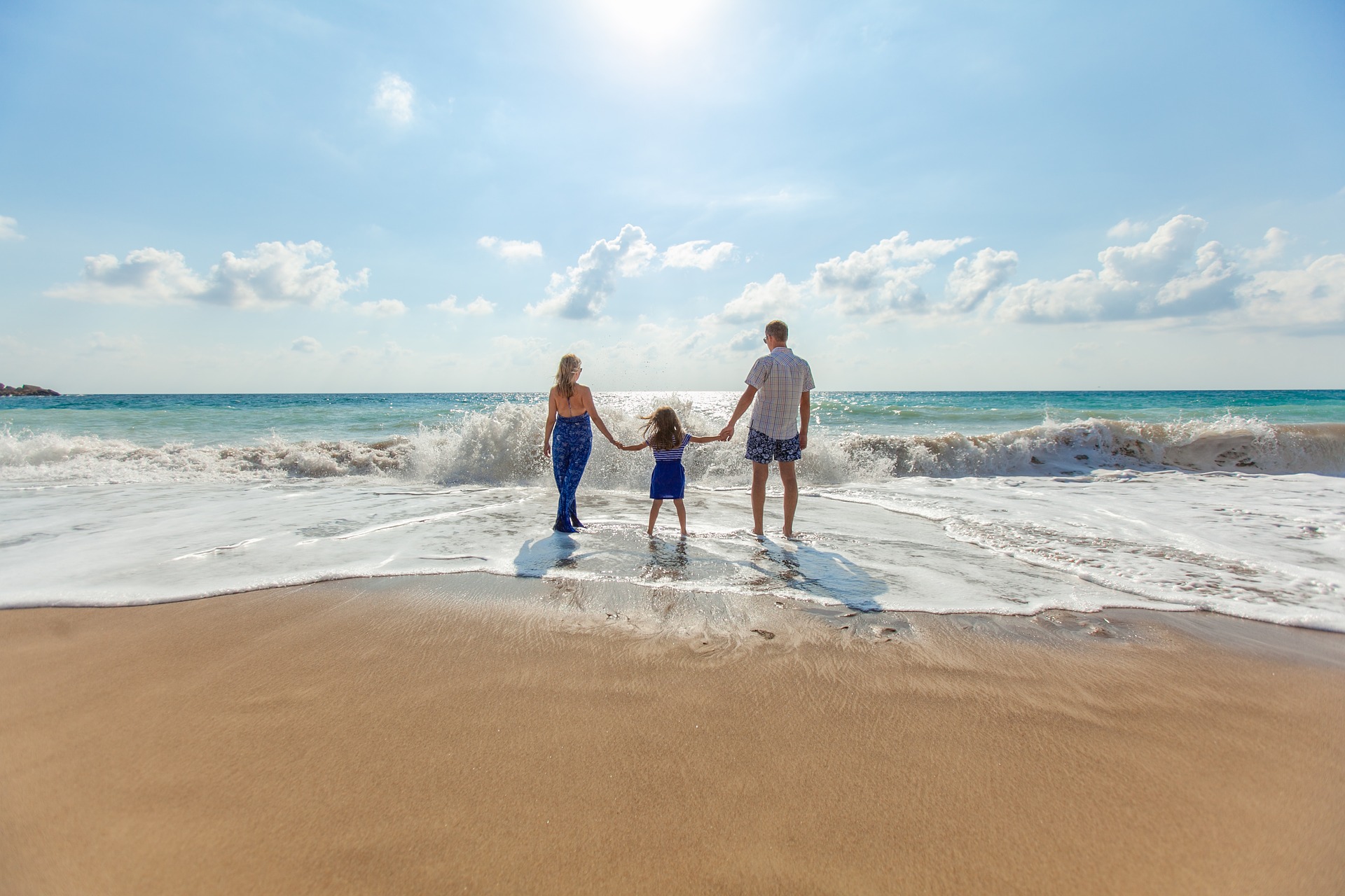 Un bambino tenuto per mano dalla mamma e dal papà. Guardano il mare sul bagnasciuga mentre un'onda bagna i loro piedi